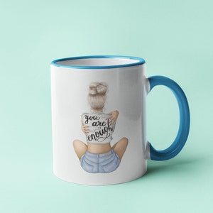 K&ouml;nnte beinhalten: Wei&szlig;e Keramiktasse mit blauem Rand und Henkel. Die Tasse zeigt eine Grafik einer Frau, die ihre Arme um sich selbst schlingt, und den Text "you are enough".