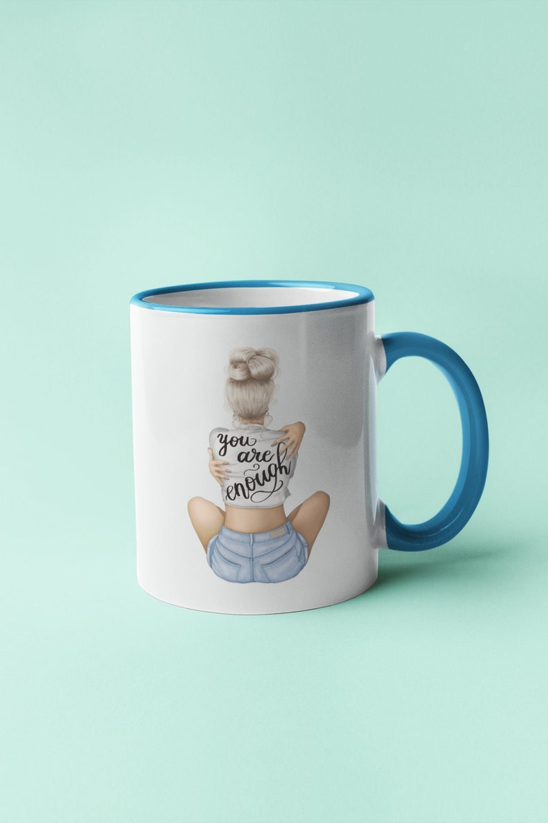 K&ouml;nnte beinhalten: Wei&szlig;e Keramiktasse mit blauem Rand und Henkel. Die Tasse zeigt eine Grafik einer Frau, die ihre Arme um sich selbst schlingt, und den Text "you are enough".
