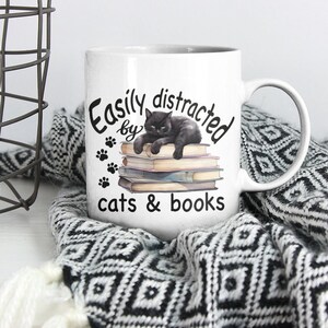 May include: White ceramic mug with a black cat illustration lying on a stack of books. The mug has the text "Easily distracted by cats & books" printed on it.