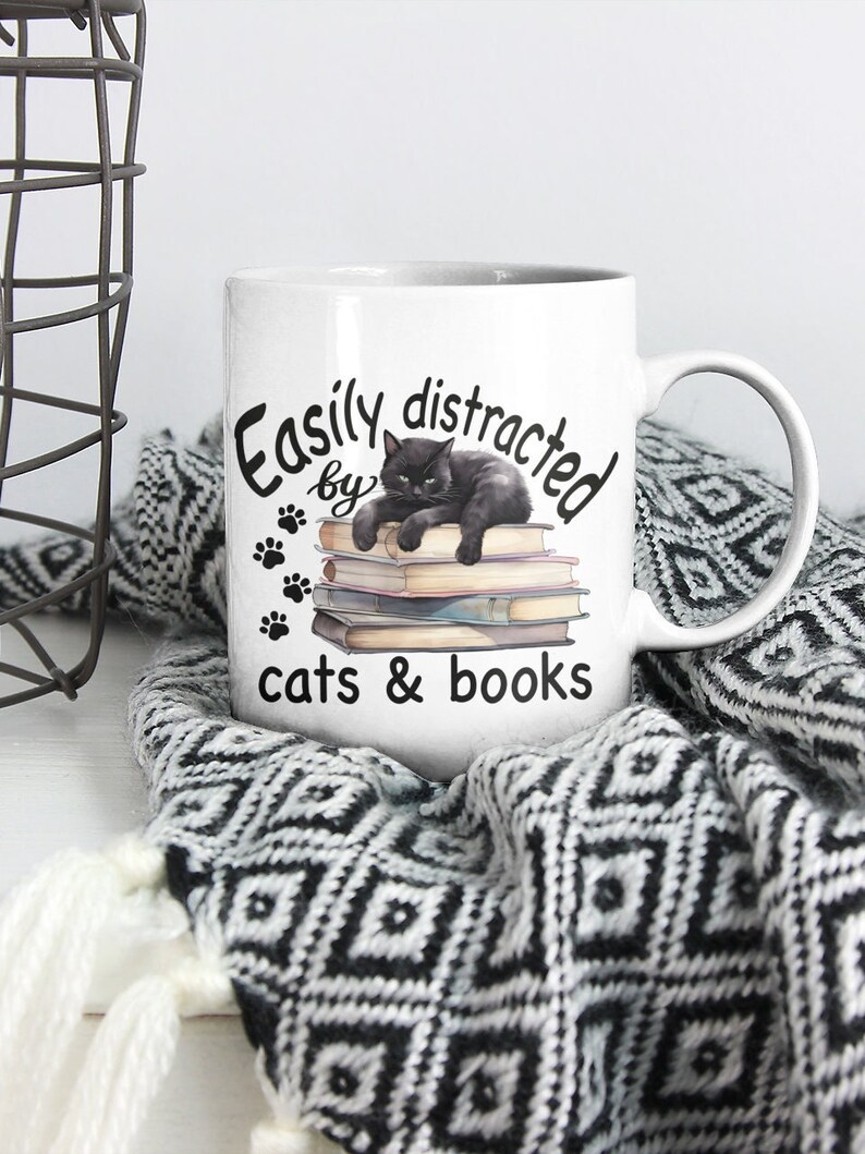 May include: White ceramic mug with a black cat illustration lying on a stack of books. The mug has the text "Easily distracted by cats & books" printed on it.