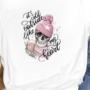 May include: White t-shirt featuring a skull wearing a pink beanie and scarf, holding a coffee cup. The shirt has the text "It's cold outside like my heart" in a script font. The design is a whimsical, winter-themed graphic.