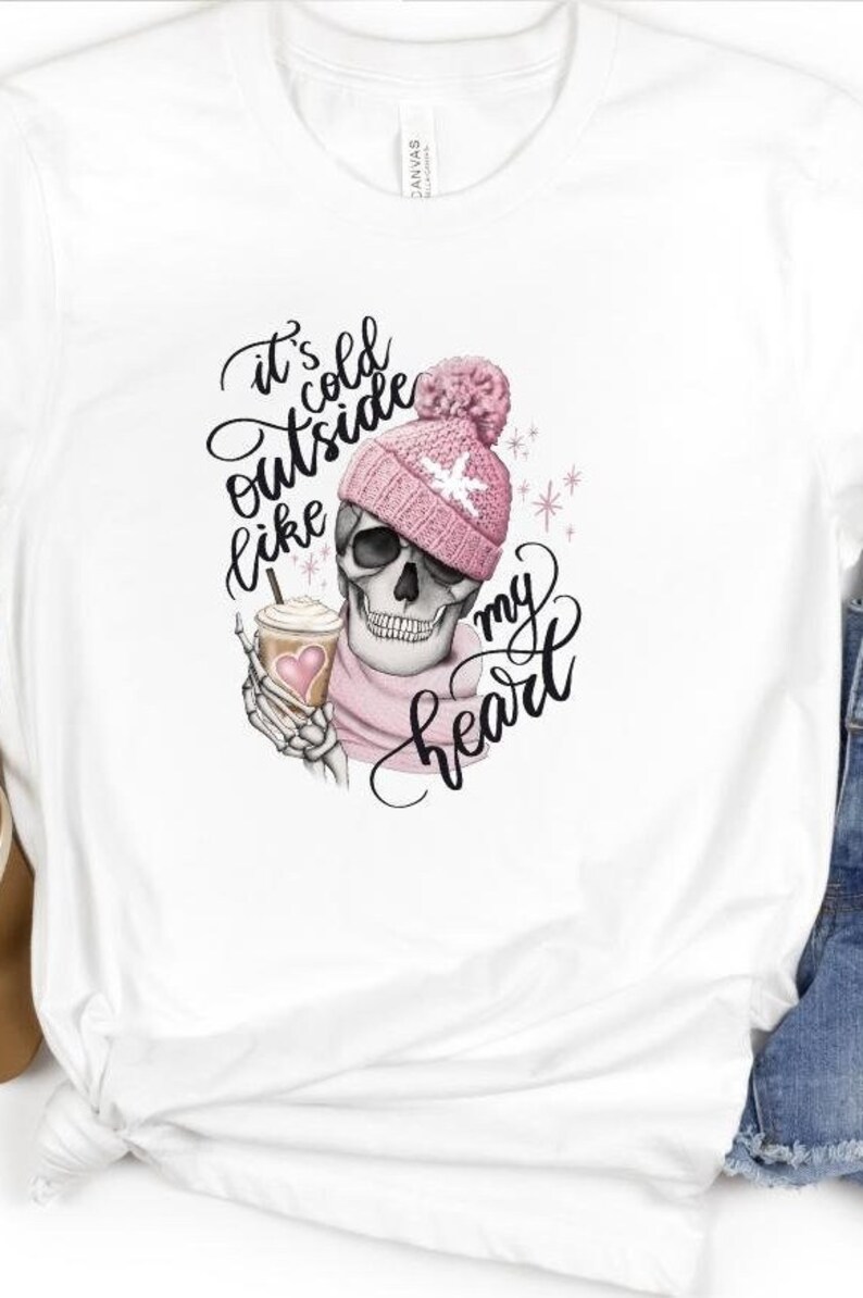 May include: White t-shirt featuring a skull wearing a pink beanie and scarf, holding a coffee cup. The shirt has the text "It's cold outside like my heart" in a script font. The design is a whimsical, winter-themed graphic.