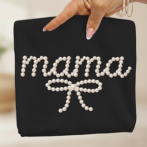 May include: Black tote bag with the word "mama" spelled out in white pearls, with a pearl bow underneath. The bag is held up by a hand with a French manicure and a pearl bracelet.