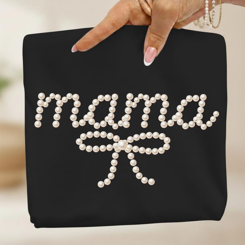 May include: Black tote bag with the word "mama" spelled out in white pearls, with a pearl bow underneath. The bag is held up by a hand with a French manicure and a pearl bracelet.