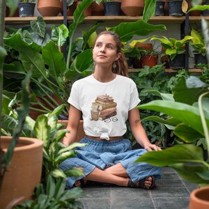 以下が含まれることがあります： 本、コーヒーカップ、眼鏡が描かれた白いTシャツ。デザインは茶色とベージュの色合いです。青いズボンを履いた人が、庭で足を組んで座っています。
