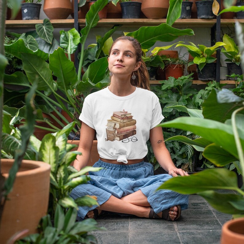 以下が含まれることがあります： 本、コーヒーカップ、眼鏡が描かれた白いTシャツ。デザインは茶色とベージュの色合いです。青いズボンを履いた人が、庭で足を組んで座っています。
