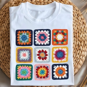 May include: White t-shirt featuring a colorful, square crochet design. The design is composed of nine individual squares, each with a unique pattern and color scheme, including shades of blue, pink, orange, and yellow. The shirt is laid flat on a woven surface.