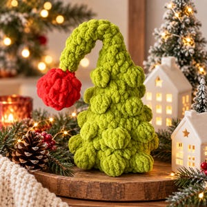 Op de afbeelding: Een gehaakte kerstboomdecoratie gemaakt van limoengroen garen met een textuurrijk, bobbelig oppervlak. De boom heeft een gebogen bovendeel dat naar beneden buigt en eindigt in een felrode, bolvormige pompon aan de uiterste punt.