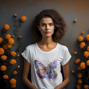 Puede incluir: Una camiseta blanca con un dise&ntilde;o de mariposa y flores coloridas. La mariposa es naranja, morada y azul, y las flores son moradas, azules y verdes. La camiseta es de estilo casual con mangas cortas y cuello redondo.