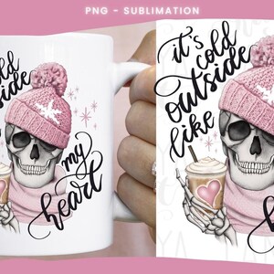 May include: White ceramic mug featuring a skull wearing a pink knit hat and scarf, holding a coffee cup with a heart. The mug has the text "It's cold outside like my heart" in black script.