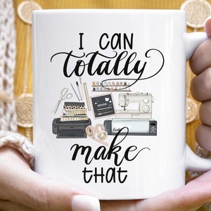 Może przedstawiać: Biały kubek ceramiczny z napisem "I can totally make that" w czarnym, ozdobnym pismie. Kubek zawiera grafikę z r&oacute;żnymi przyborami do rękodzieła, w tym maszyną do szycia, nożyczkami, nicią i drukarką.