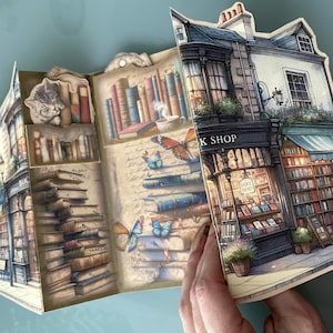 May include: A pop-up book with a whimsical illustration of a bookstore with stacks of books, a sleeping cat, and butterflies. The book is open to reveal the interior of the shop, which is filled with bookshelves and a sign that reads "K Shop".