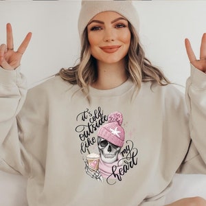 May include: Beige sweatshirt with a graphic of a skull wearing a pink beanie and scarf, holding a coffee. The text reads "It's cold outside like my heart".