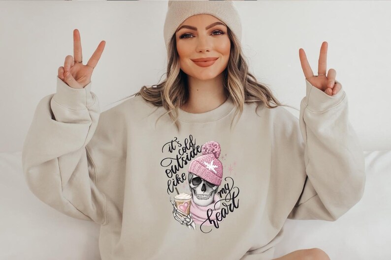 May include: Beige sweatshirt with a graphic of a skull wearing a pink beanie and scarf, holding a coffee. The text reads "It's cold outside like my heart".