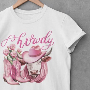 May include: White t-shirt featuring a pink cow wearing a pink cowboy hat and pink boots. The text "Howdy" is written in pink cursive above the cow.
