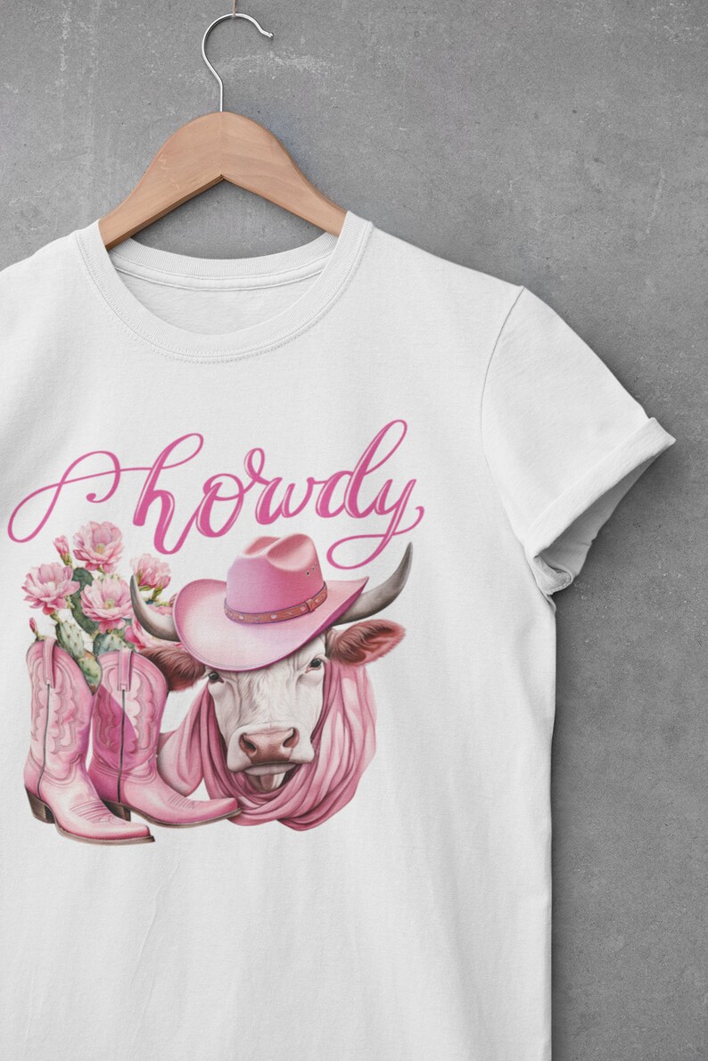 May include: White t-shirt featuring a pink cow wearing a pink cowboy hat and pink boots. The text "Howdy" is written in pink cursive above the cow.