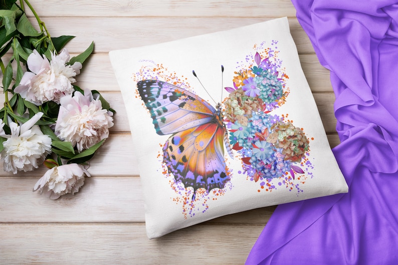 Puede incluir: Una almohada blanca con una mariposa colorida y un dise&ntilde;o floral. La mariposa es naranja, morada y azul con marcas negras. Las flores son azules, moradas y naranjas.