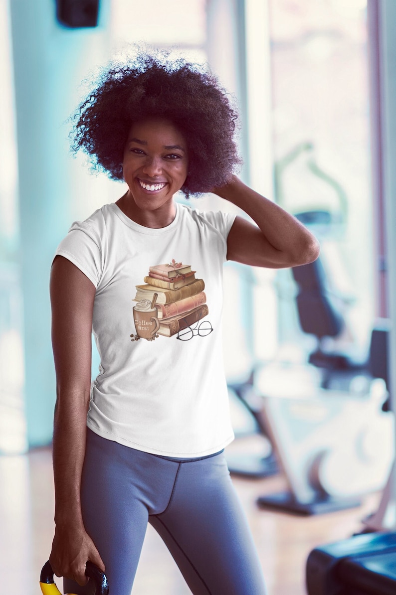 以下が含まれることがあります： 白いTシャツには、本が積み重ねられたデザイン、"Coffee Here"の文字が入ったコーヒーカップ、メガネが描かれています。デザインは茶色とベージュの色合いです。人物はグレーのレギンスを着用しています。