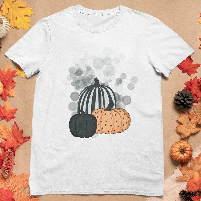 Puede incluir: Camiseta blanca con un dise&ntilde;o de tres calabazas. Una es negra, otra naranja con lunares negros y la tercera a rayas blancas y negras. El dise&ntilde;o incluye detalles de burbujas grises, perfecto para el oto&ntilde;o.