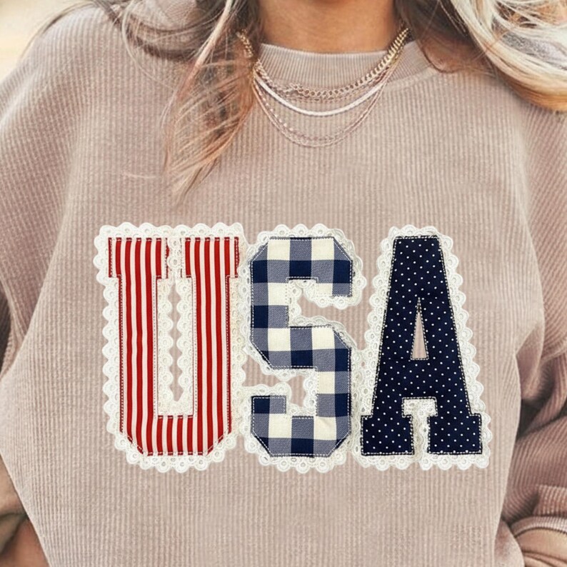 May include: A tan-colored sweatshirt with the word "USA" appliqued on the front. The letters are in red and white stripes, blue and white gingham, and navy blue with white polka dots, all outlined with white lace.