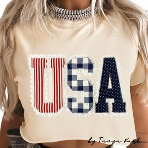 May include: Cream-colored crop top with the word "USA" in red and white striped, blue gingham, and navy blue polka dot letters. The letters are outlined with white lace. The text "by Tanya Kart" is at the bottom.