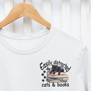 May include: White t-shirt with a black cat sitting on a stack of books. The text reads "Easily distracted by cats & books".