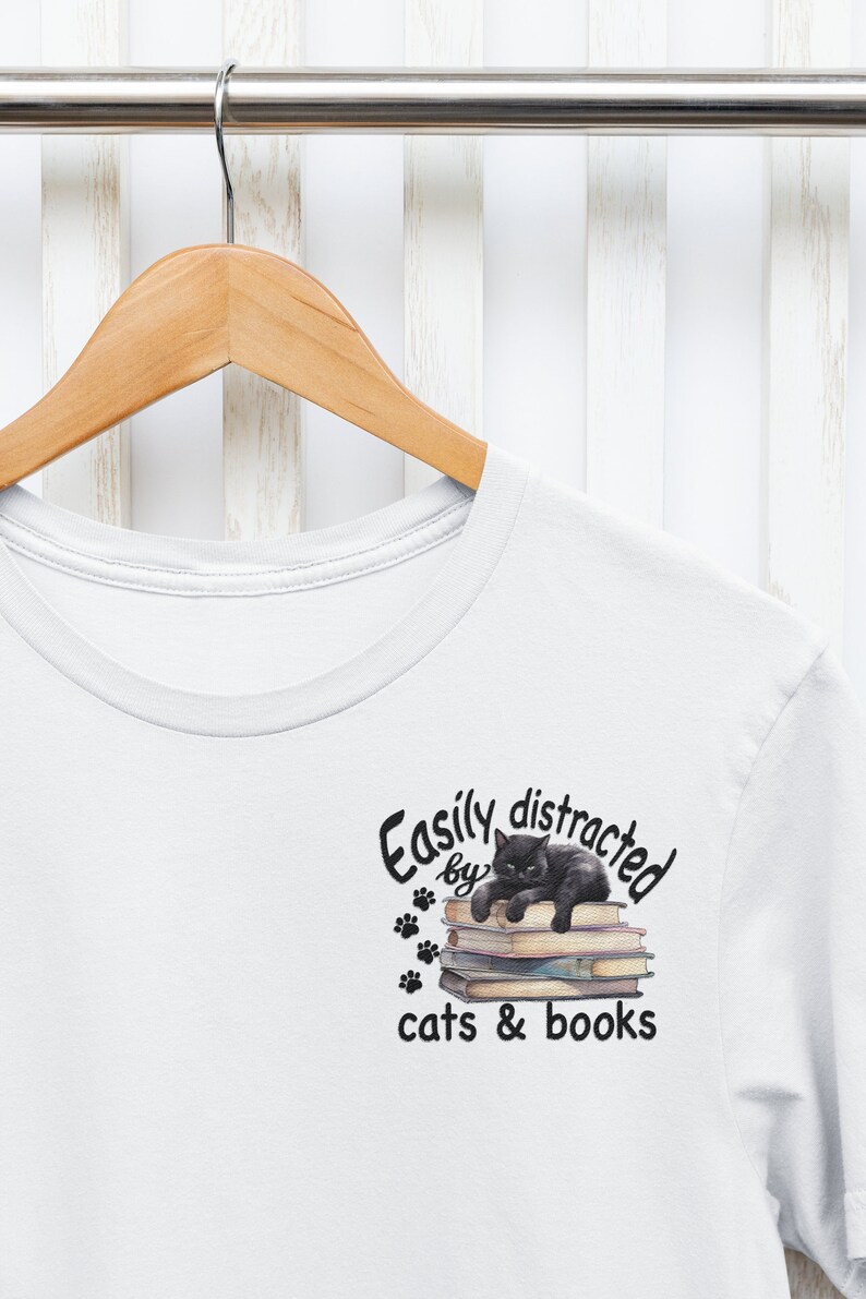 May include: White t-shirt with a black cat sitting on a stack of books. The text reads "Easily distracted by cats & books".