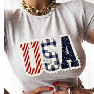 May include: A light gray t-shirt with the word "USA" appliqued on the front. The letters are in red and white stripes, blue gingham, and navy blue with white polka dots, all trimmed with white lace.