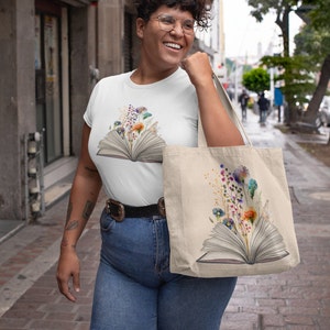 Pu&ograve; includere: Una maglietta bianca con un design floreale colorato su un libro aperto. La maglietta &egrave; indossata da una persona con i capelli ricci e gli occhiali. La persona porta anche una borsa tote con lo stesso design della maglietta.