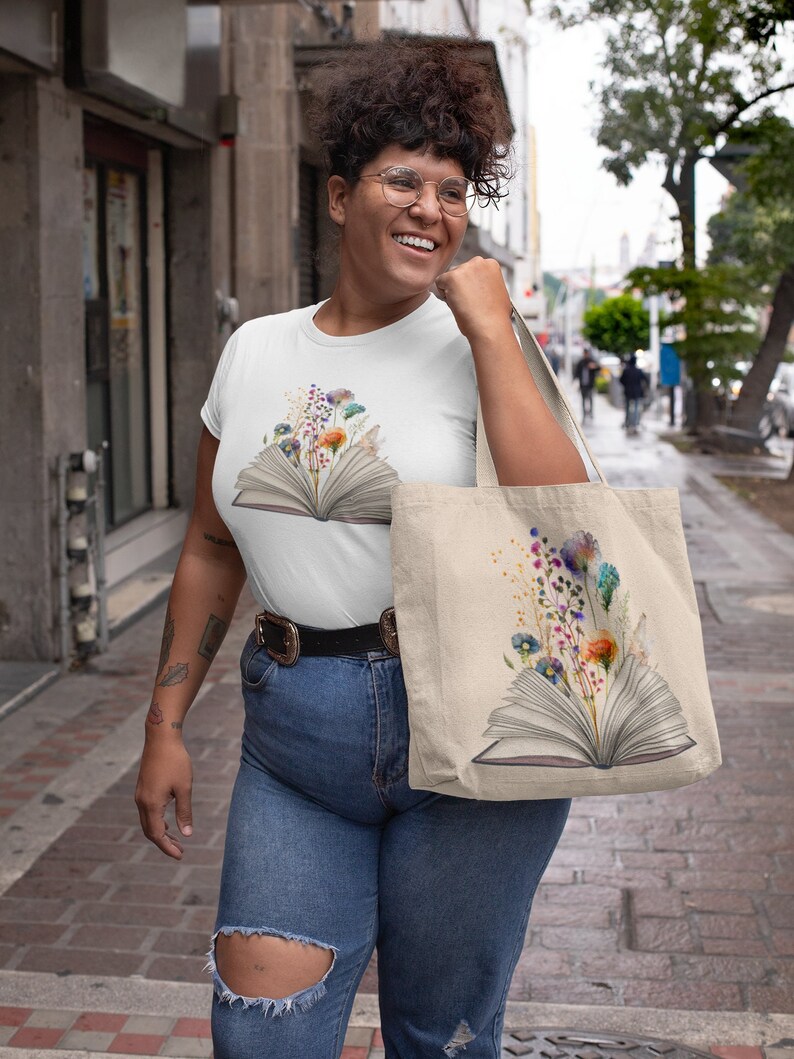 Pu&ograve; includere: Una maglietta bianca con un design floreale colorato su un libro aperto. La maglietta &egrave; indossata da una persona con i capelli ricci e gli occhiali. La persona porta anche una borsa tote con lo stesso design della maglietta.