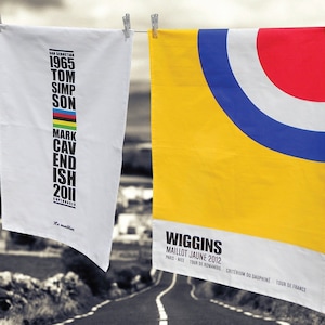 Puede incluir: Dos toallas de cocina blancas colgadas de un tendedero. La toalla de la izquierda presenta un diseño en blanco y negro con los nombres de los ganadores anteriores del Tour de Francia, incluidos Tom Simpson y Mark Cavendish. La toalla de la derecha presenta un diseño amarillo, azul, rojo y blanco con el texto "Wiggins Maillot Jaune 2012".