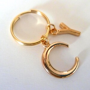 May include: A gold-colored keychain with a circular ring, a crescent moon charm, and a letter "Y" charm. The keychain is on a white surface. The moon charm is a curved shape, and the letter "Y" is a simple, block-style design.