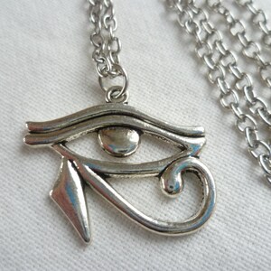 May include: Silver-toned pendant necklace featuring the Eye of Horus symbol. The pendant is detailed with an eye shape and a curved design, suspended from a linked chain. The necklace is displayed on a white surface.