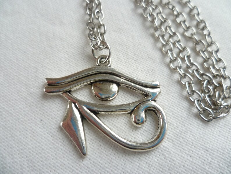 May include: Silver-toned pendant necklace featuring the Eye of Horus symbol. The pendant is detailed with an eye shape and a curved design, suspended from a linked chain. The necklace is displayed on a white surface.