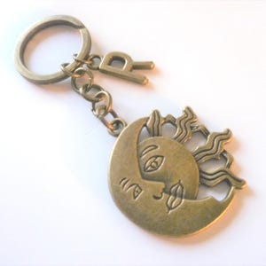 May include: Antique brass-coloured keyring featuring a sun and moon design. The sun and moon are intertwined, with the letter "R" attached. The keyring has a split ring and chain.