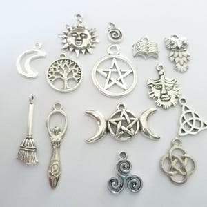 Op de afbeelding: Een verzameling zilverkleurige bedels met diverse mystieke en Wicca-symbolen. Bedels zijn onder meer een halve maan, een zonnegelaat, een levensboom, een pentagram, een uil, een boek, een bezem, een godinnenfiguur en een drievoudige maan.
