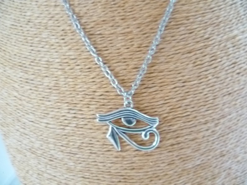 May include: Silver-toned necklace featuring an Eye of Horus pendant. The pendant is a stylized eye with intricate details, suspended from a delicate chain. The necklace is displayed against a neutral background.