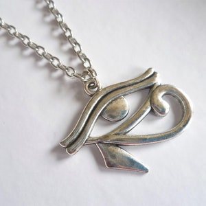May include: Silver-toned metal pendant necklace featuring the Eye of Horus symbol. The pendant is detailed with curved lines and a teardrop shape, suspended from a delicate chain. The necklace is displayed on a white surface.