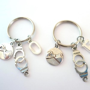 May include: Two silver keychains with a pinkie promise charm, handcuffs with the word "Freedom" on them, and a letter charm. One keychain has the letter "E" charm and the other has a silver oval charm.