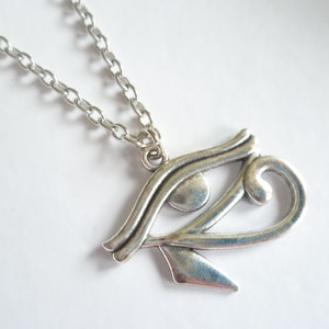 May include: Silver-toned pendant necklace featuring the Eye of Horus symbol. The pendant is a stylized representation of an eye, with intricate details and a polished finish. The chain is a simple, linked design.