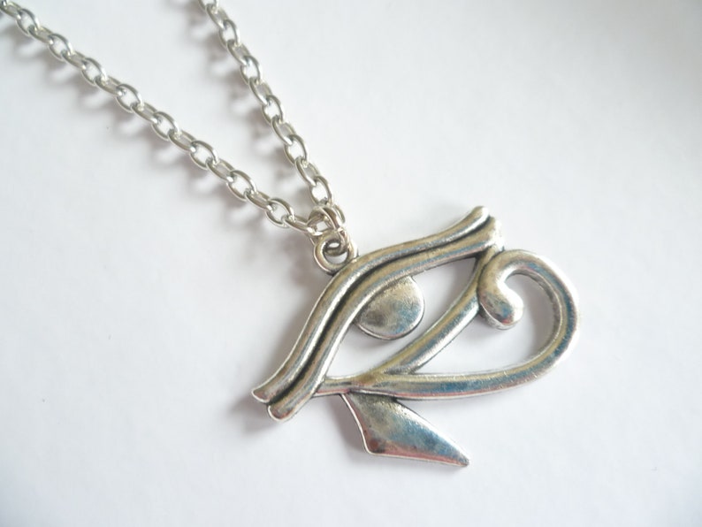 May include: Silver-toned pendant necklace featuring the Eye of Horus symbol. The pendant is a stylized representation of an eye, with intricate details and a polished finish. The chain is a simple, linked design.