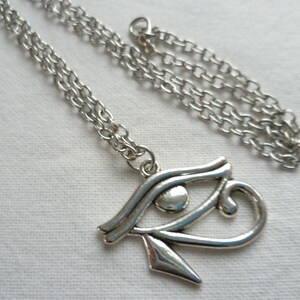 May include: Silver-toned necklace featuring an Eye of Horus pendant. The pendant is a stylized eye symbol with intricate details, suspended from a delicate chain. The necklace is displayed on a white surface, highlighting the metallic sheen of the jewelry.