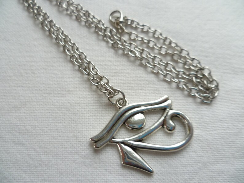 May include: Silver-toned necklace featuring an Eye of Horus pendant. The pendant is a stylized eye symbol with intricate details, suspended from a delicate chain. The necklace is displayed on a white surface, highlighting the metallic sheen of the jewelry.