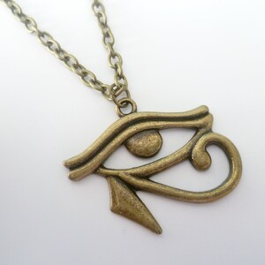 May include: A bronze-colored pendant necklace featuring the Eye of Horus symbol. The pendant is detailed with curved lines and a teardrop shape, suspended from a matching chain. The necklace is displayed against a white background.