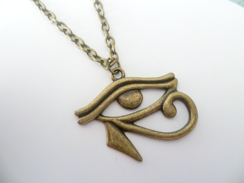 May include: A bronze-colored pendant necklace featuring the Eye of Horus symbol. The pendant is detailed with curved lines and a teardrop shape, suspended from a matching chain. The necklace is displayed against a white background.