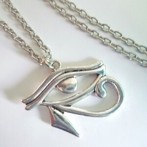 May include: A silver-colored necklace featuring an Eye of Horus pendant. The pendant is detailed with curved lines and a teardrop-shaped eye. The chain is a simple link style, and the necklace is displayed on a white surface.