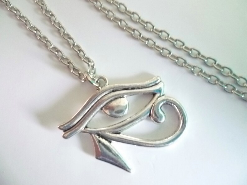 May include: A silver-colored necklace featuring an Eye of Horus pendant. The pendant is detailed with curved lines and a teardrop-shaped eye. The chain is a simple link style, and the necklace is displayed on a white surface.