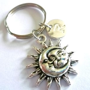 May include: Silver-coloured keyring with a sun and moon charm. The sun has pointed rays and a smiling face, with a crescent moon overlay. A small, round charm with the letter "R" is also attached. The keyring is on a silver ring.