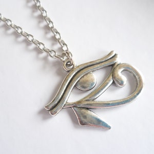 May include: A silver-colored pendant necklace featuring the Eye of Horus symbol. The pendant is detailed with curved lines and a teardrop shape, suspended from a linked chain. The necklace is displayed against a white background.