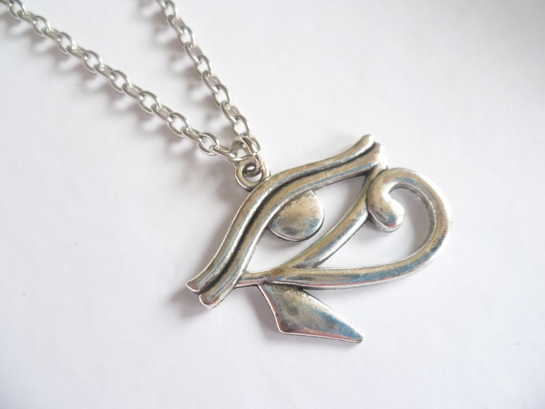 May include: A silver-colored pendant necklace featuring the Eye of Horus symbol. The pendant is detailed with curved lines and a teardrop shape, suspended from a linked chain. The necklace is displayed against a white background.
